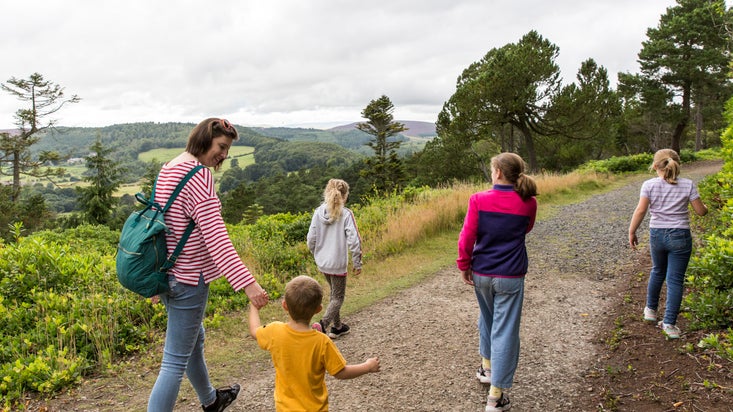 An adult with four children on a visit at Cragside. They are walking along a rugged path with views of the Northumberland Hills in the distance.